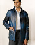 Male model, half front shot, wearing our new Poseidon style, a two-tone black and blue chore jacket, unbuttoned with one hand in one of the pockets, wearing a white dress shirt, a belt and dark pants on a white background.