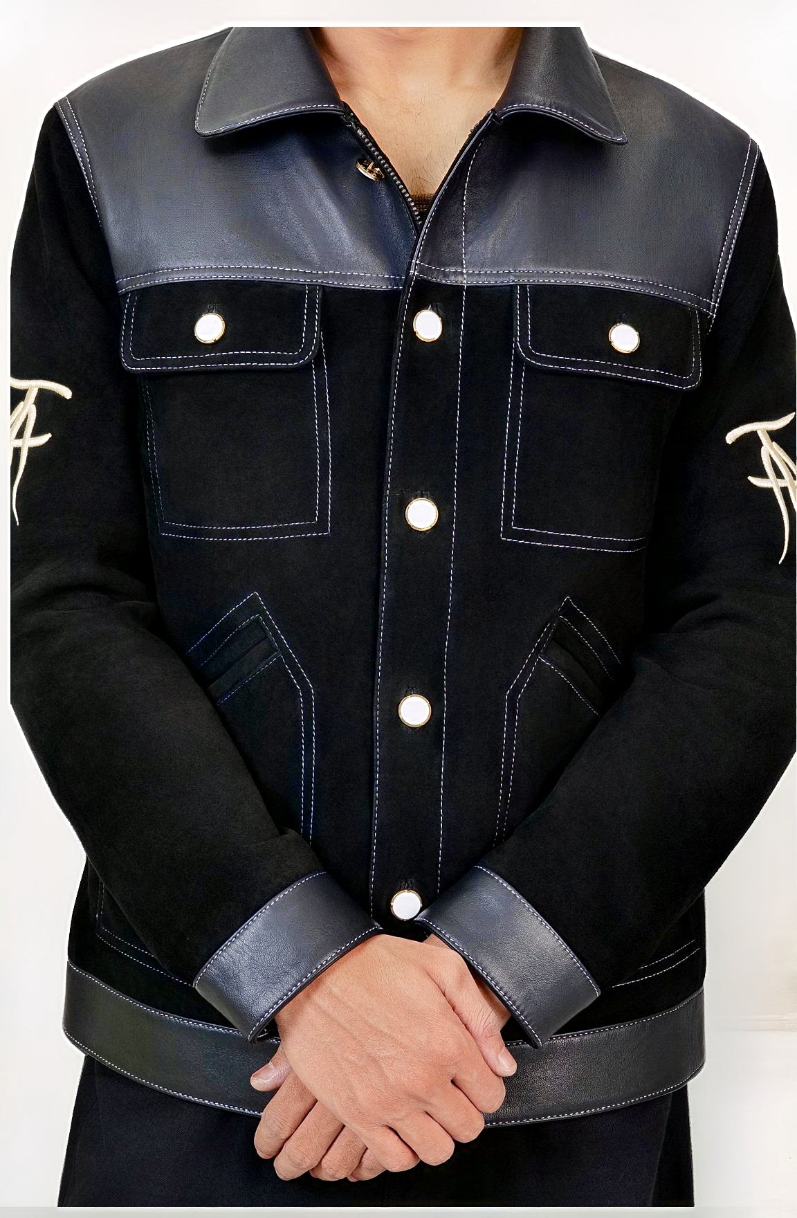 Male model detail shot, wearing our new Ares style, a Black suede/nubuck trucker jacket with black leather collar, upper front/chest, cuff and base trim showcasing mid- sleeve "T" and "A" embroidery detail, white contrast stitching and white body and top pocket button closures, buttoned up with white button closures, wearing dark grey cropped pants on a white background.