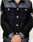 Male model detail shot, wearing our new Ares style, a Black suede/nubuck trucker jacket with black leather collar, upper front/chest, cuff and base trim showcasing mid- sleeve "T" and "A" embroidery detail, white contrast stitching and white body and top pocket button closures, buttoned up with white button closures, wearing dark grey cropped pants on a white background.