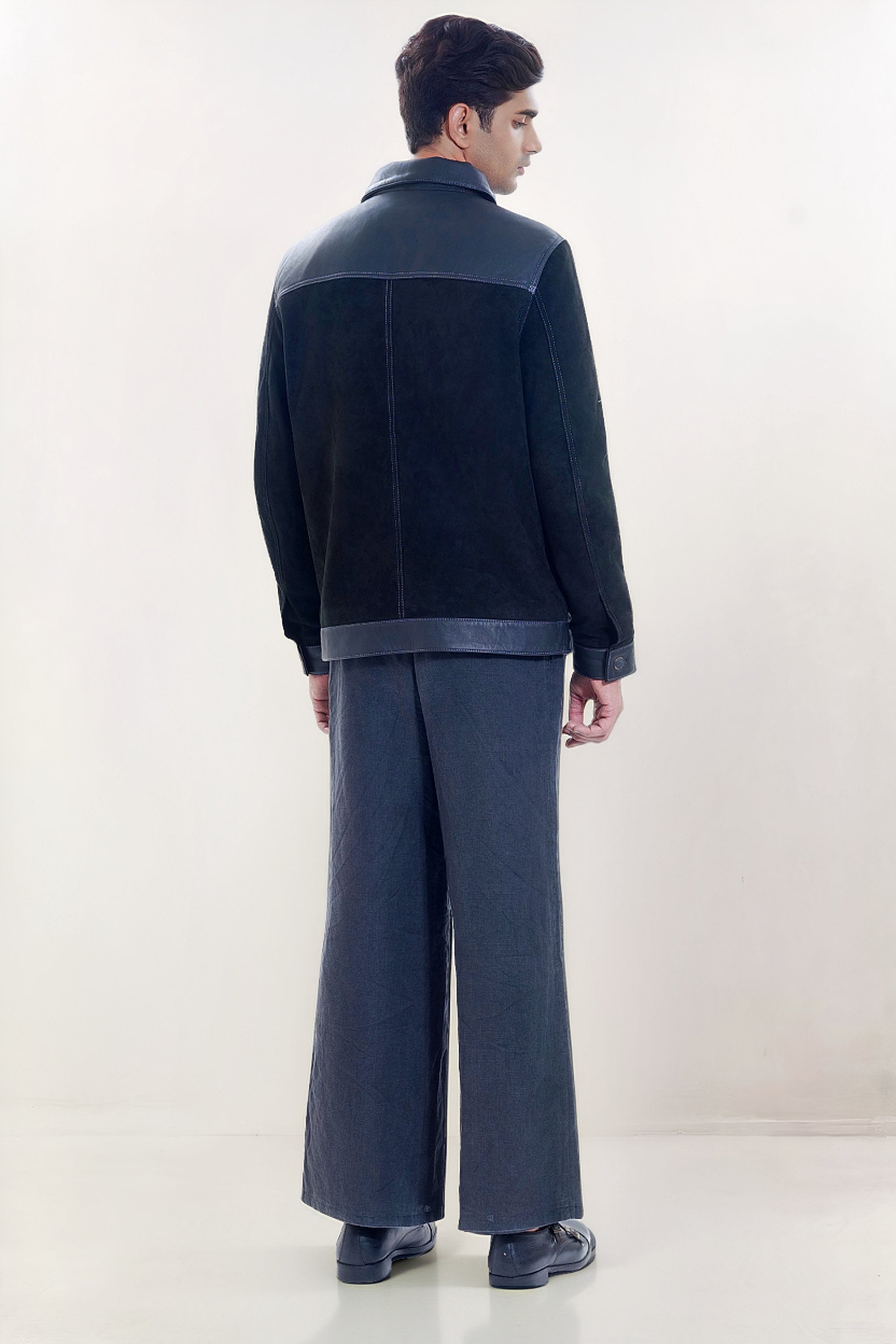 Male model full back shot, wearing our new Ares style, a Black suede/nubuck trucker jacket with black leather collar, upper back, cuff and base trim showcasing button closure cuffs and dark grey cropped pants with loafer dress shoes on a white background.