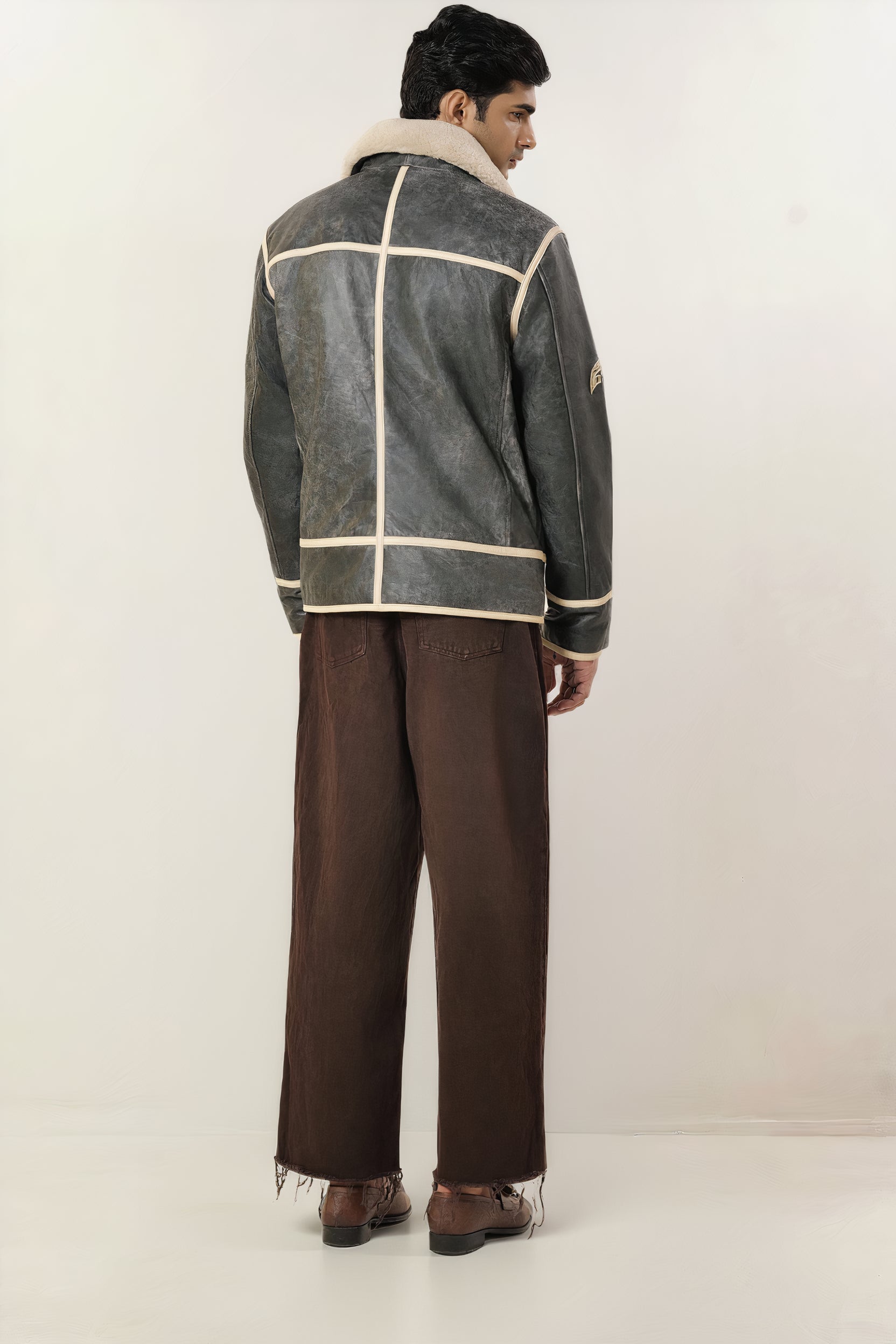Male model full back shot, wearing our new Icarus style, a vintage-looking weathering grey colour flight jacket showcasing embroidery detail, off-white leather strips, brown pants, and dress loafers on a white background.