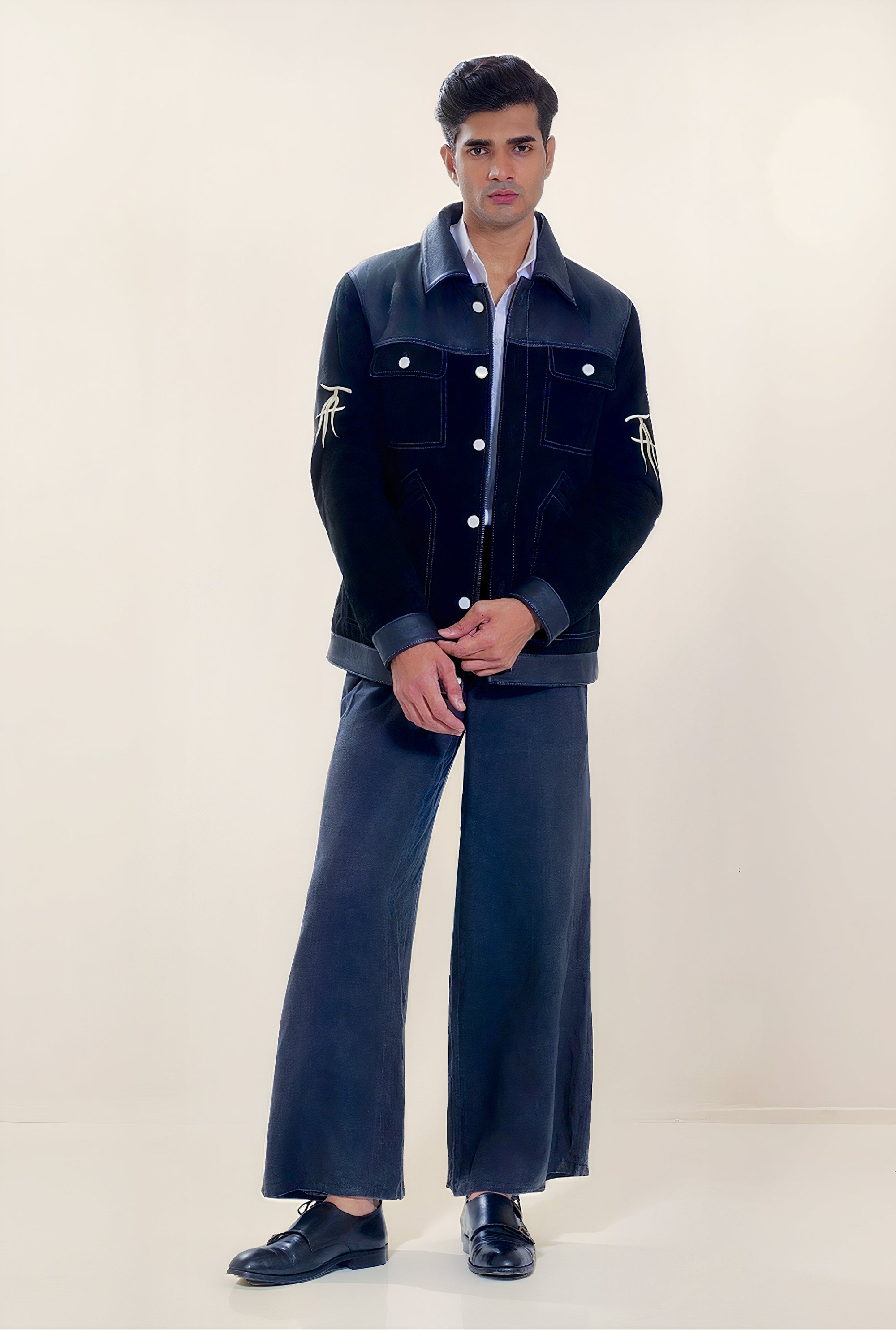 Male model full front shot, wearing our new Ares style, a Black suede/nubuck trucker jacket with black leather collar, upper front/chest, cuff and base trim showcasing mid- sleeve "T" and "A" embroidery detail, white contrast stitching and white body and top pocket button closures,  a white dress shirt, and dark grey cropped pants with loafer dress shoes on a white background.