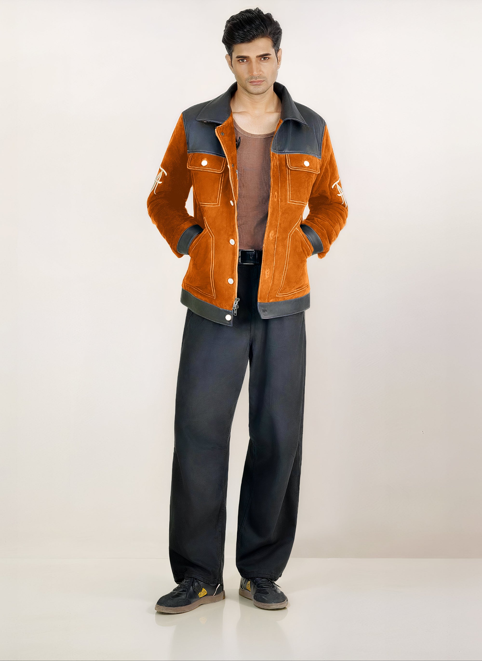 Male model full front shot, wearing our new Ares style, a Cognac suede/nubuck trucker jacket with black leather collar, upper front/chest, cuff and base trim showcasing mid- sleeve "T" and "A" embroidery detail, white contrast stitching and white body and top pocket button closures,  a white dress shirt, and dark grey cropped pants with loafer dress shoes on a white background.