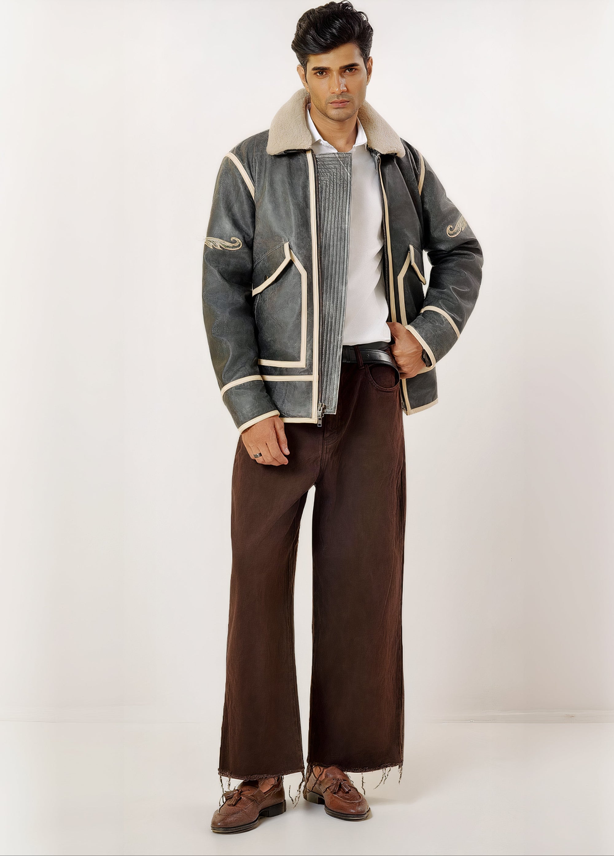 Male model full front shot, wearing our new Icarus style, a vintage-looking weathering grey colour flight jacket showcasing embroidery detail, off-white leather strips,a white shirt, brown pants, and dress loafers on a white background.