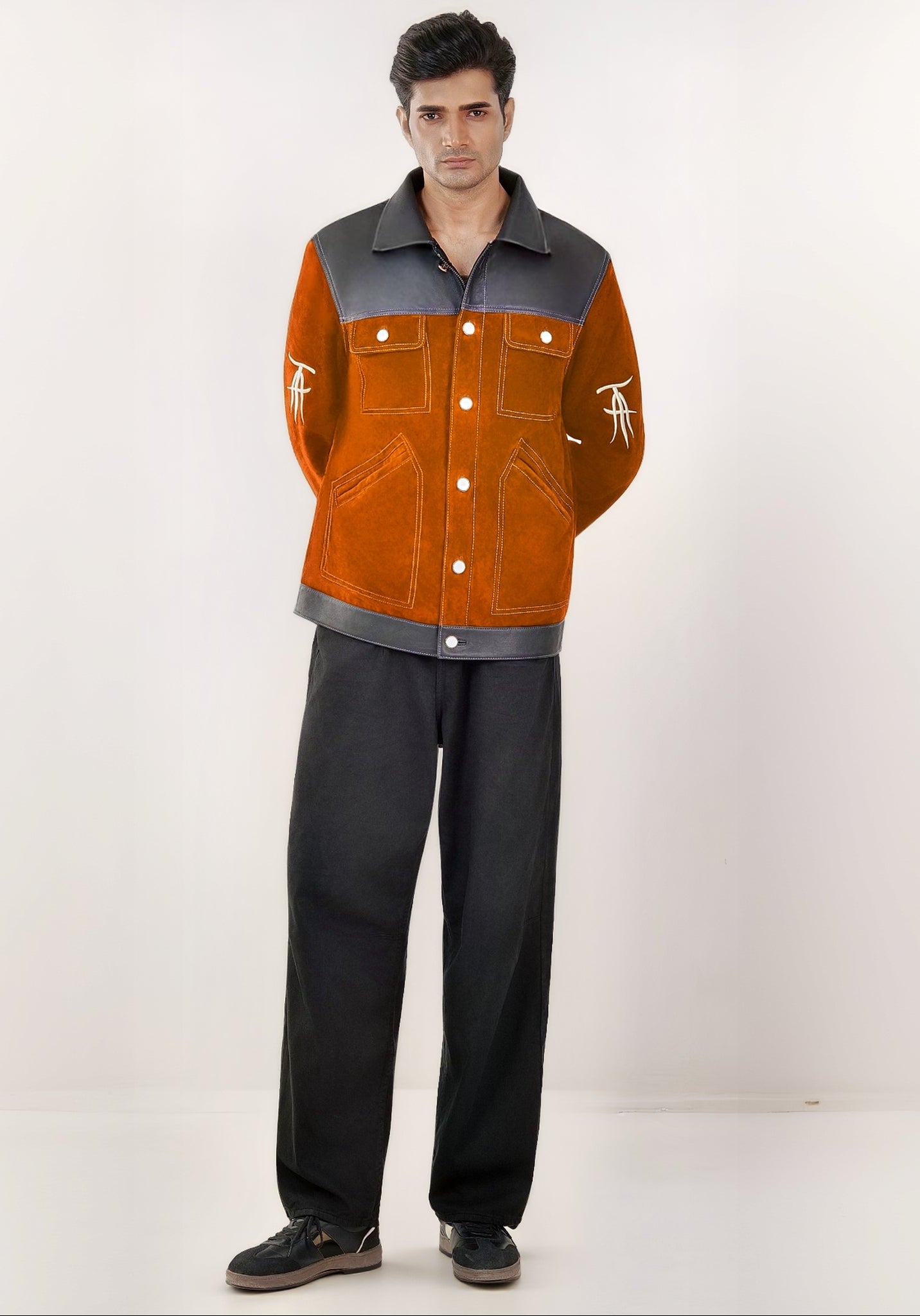 Male model full front shot, wearing our new Ares style, a Cognac suede/nubuck trucker jacket, buttoned up, with black leather collar, upper front/chest, cuff and base trim showcasing mid- sleeve &quot;T&quot; and &quot;A&quot; embroidery detail, white contrast stitching and white body and top pocket button closures,  a white dress shirt, and dark grey cropped pants with loafer dress shoes on a white background.