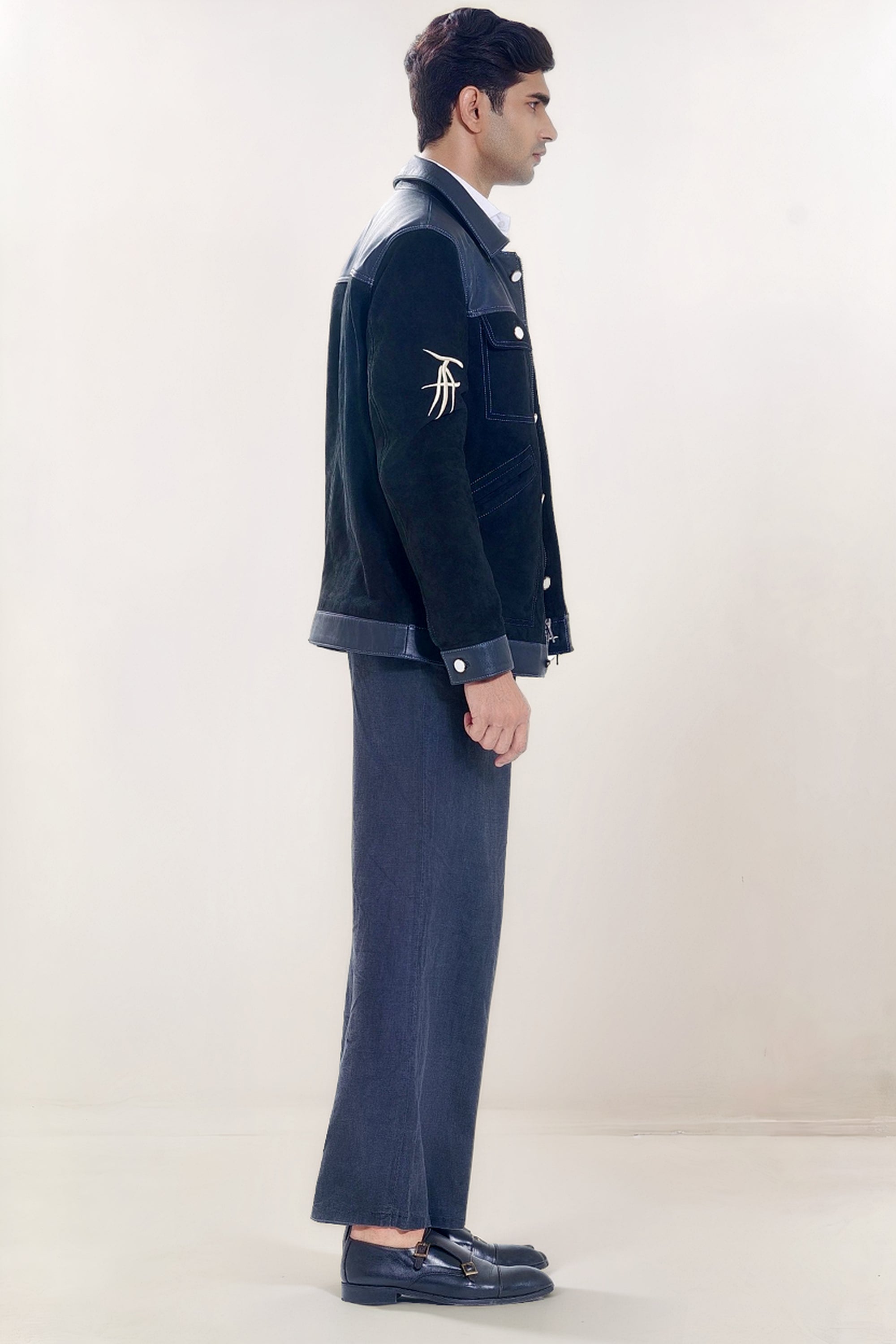 Male model full side shot, wearing our new Ares style, a Black suede/nubuck trucker jacket with black leather collar, upper front/chest, cuff and base trim showcasing mid- sleeve "T" and "A" embroidery detail, white contrast stitching and white body and top pocket button closures,  a white dress shirt, and dark grey cropped pants with loafer dress shoes on a white background.