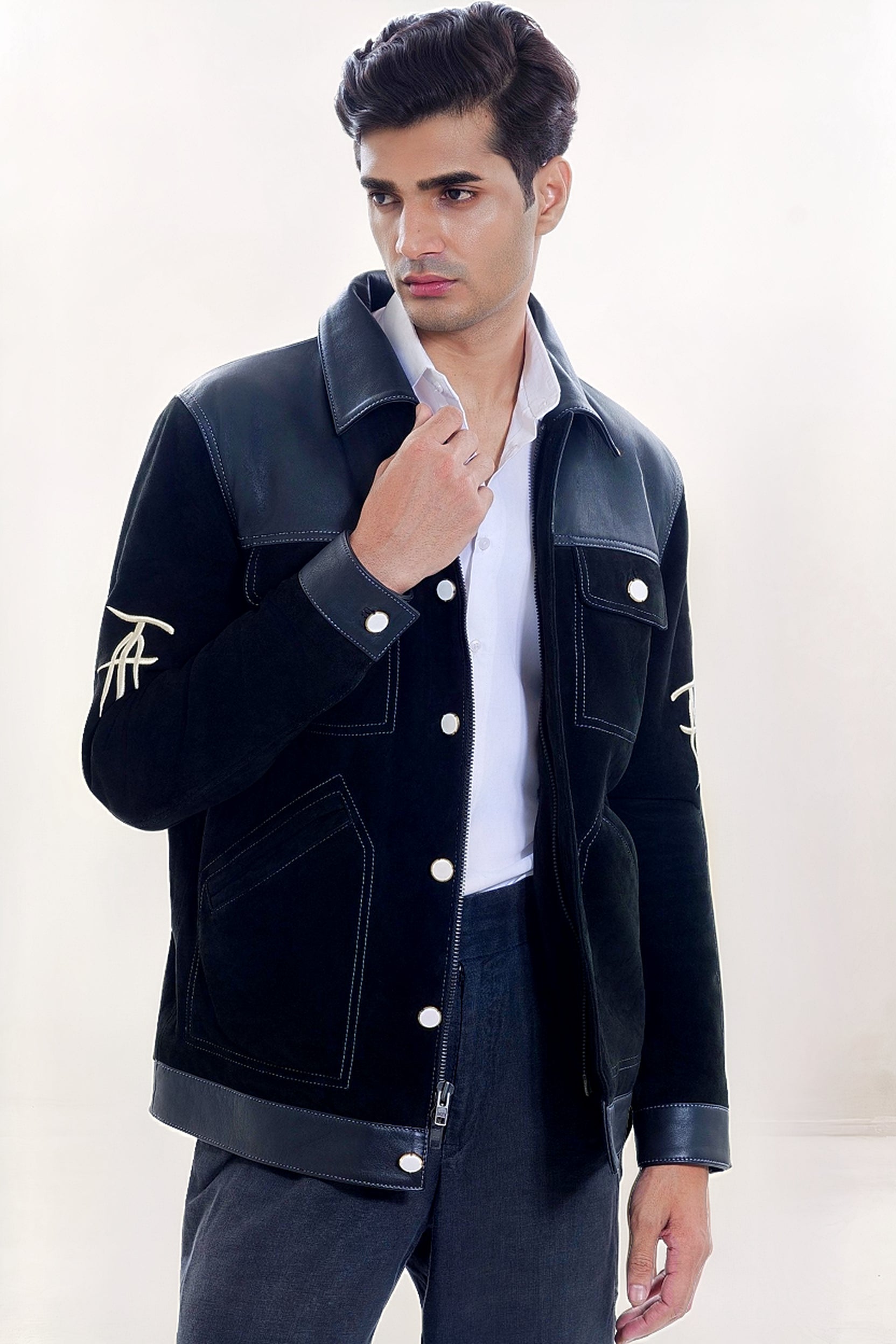 Male model half front shot, wearing our new Ares style, a Black suede/nubuck trucker jacket with black leather collar, upper front/chest, cuff and base trim showcasing mid- sleeve "T" and "A" embroidery detail, white contrast stitching and white body and top pocket button closures,  a white dress shirt, and dark grey cropped pants on a white background.