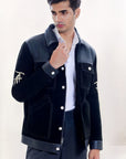 Male model half front shot, wearing our new Ares style, a Black suede/nubuck trucker jacket with black leather collar, upper front/chest, cuff and base trim showcasing mid- sleeve "T" and "A" embroidery detail, white contrast stitching and white body and top pocket button closures,  a white dress shirt, and dark grey cropped pants on a white background.
