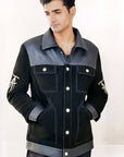 Male model half front shot, wearing our new Ares style, a Black suede/nubuck trucker jacket with black leather collar, upper front/chest, cuff and base trim showcasing mid- sleeve "T" and "A" embroidery detail, white contrast stitching and white body and top pocket button closures, buttoned up with white button closures, wearing dark grey cropped pants on a white background.