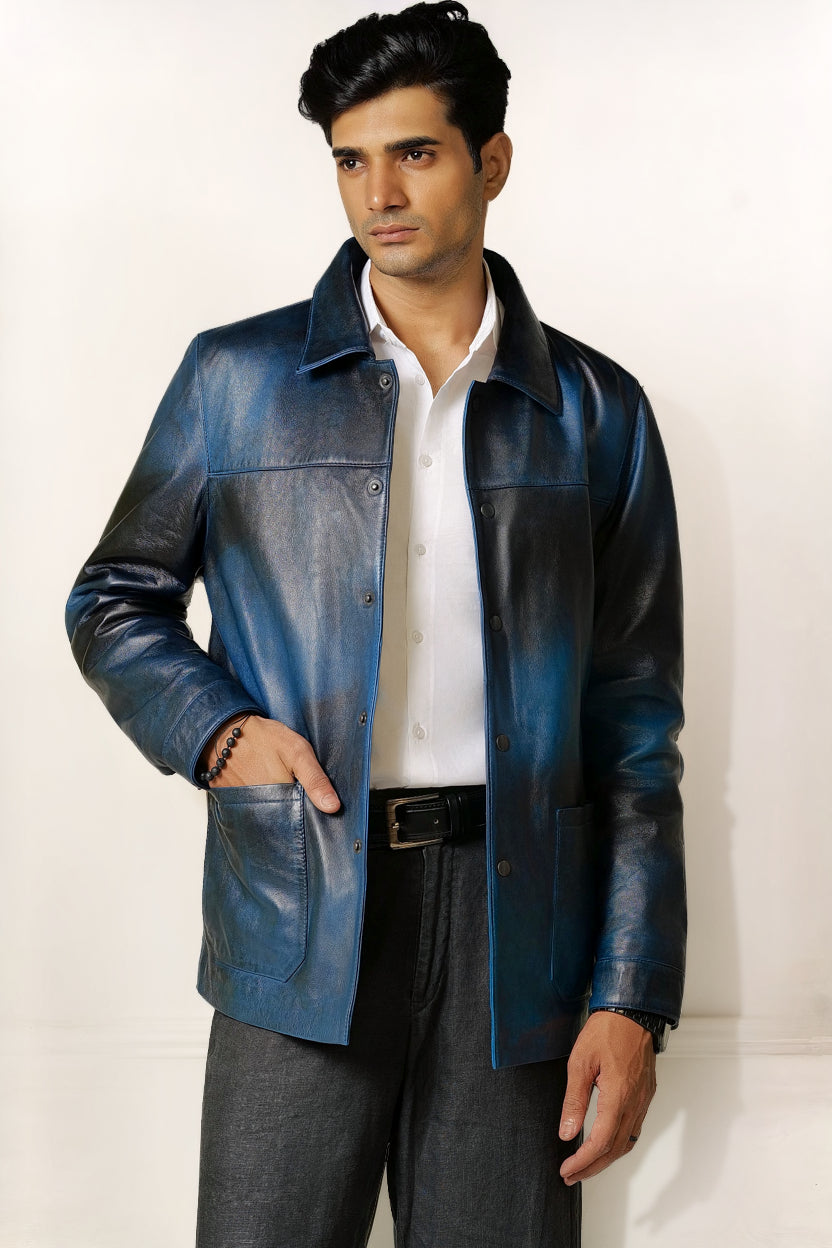 Male model, half front shot, wearing our new Poseidon style, a two-tone black and blue chore jacket, unbuttoned with one hand in one of the pockets, wearing a white dress shirt, a belt and dark pants on a white background.