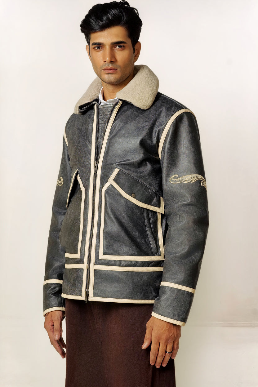 Male model, half side shot, wearing our new Icarus style, a vintage-looking weathering grey colour flight jacket, 3/4 zipped, showcasing embroidery detail, off-white leather strips, wearing brown pants, on a white background.